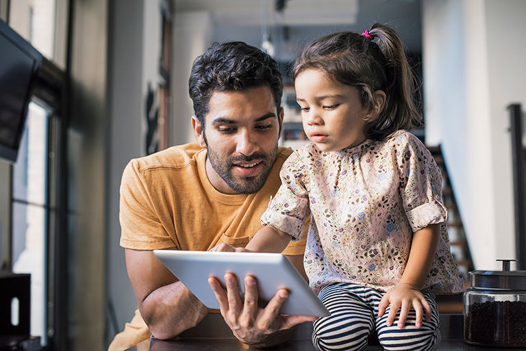 A father sitting down looking at a tablet with his young daughter.