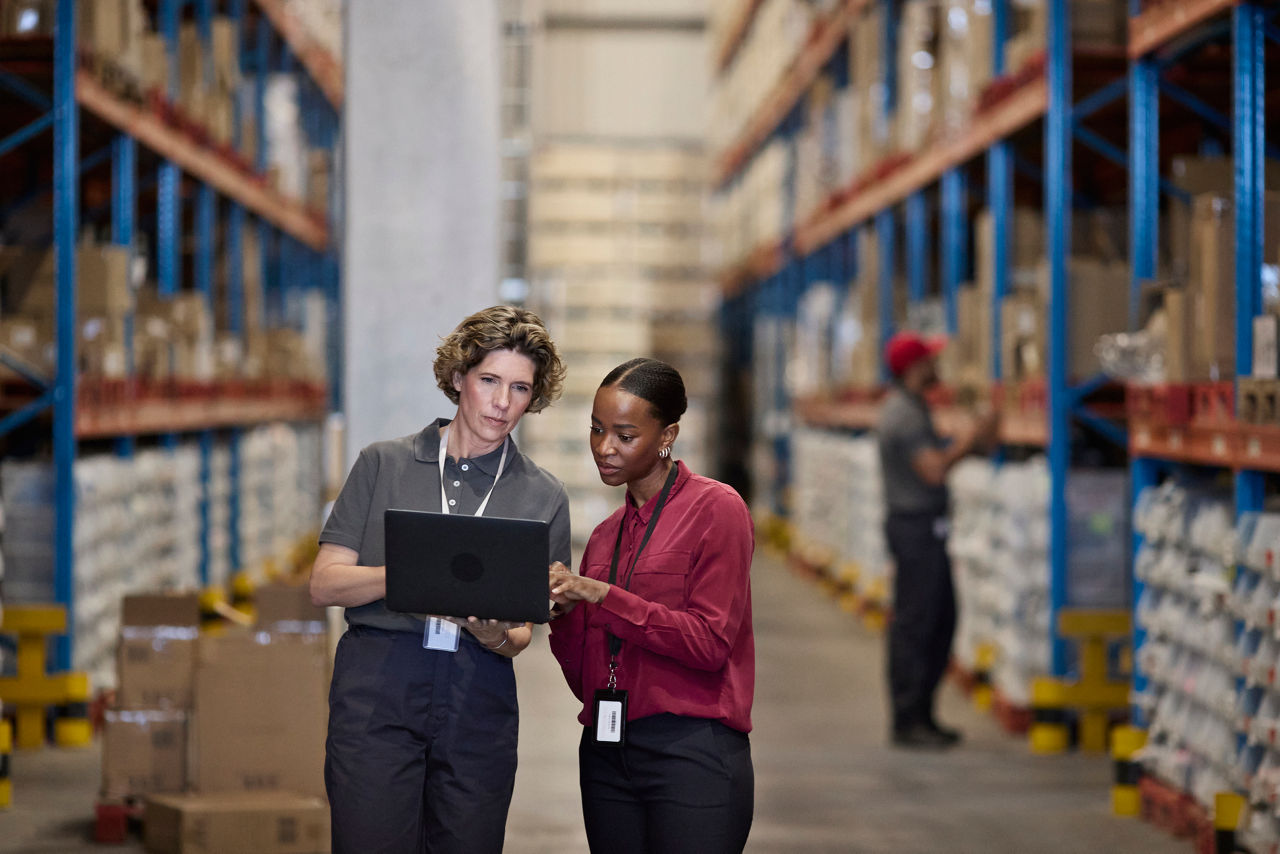 Female manager discussing with employee using laptop while standing in distribution warehouse