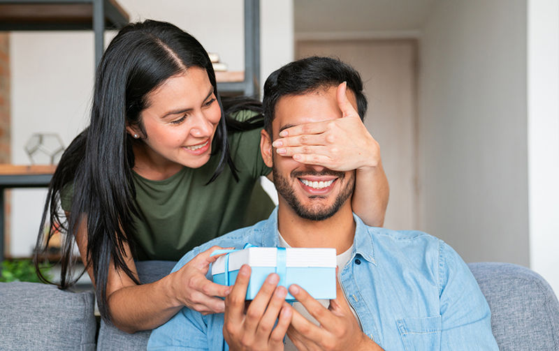 Loving Latin American woman surprising her boyfriend with a gift at home â   lifestyle concepts