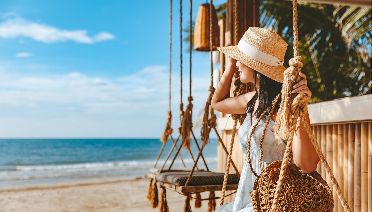 Woman with hat and dress relaxes on a swing at a beach cafe