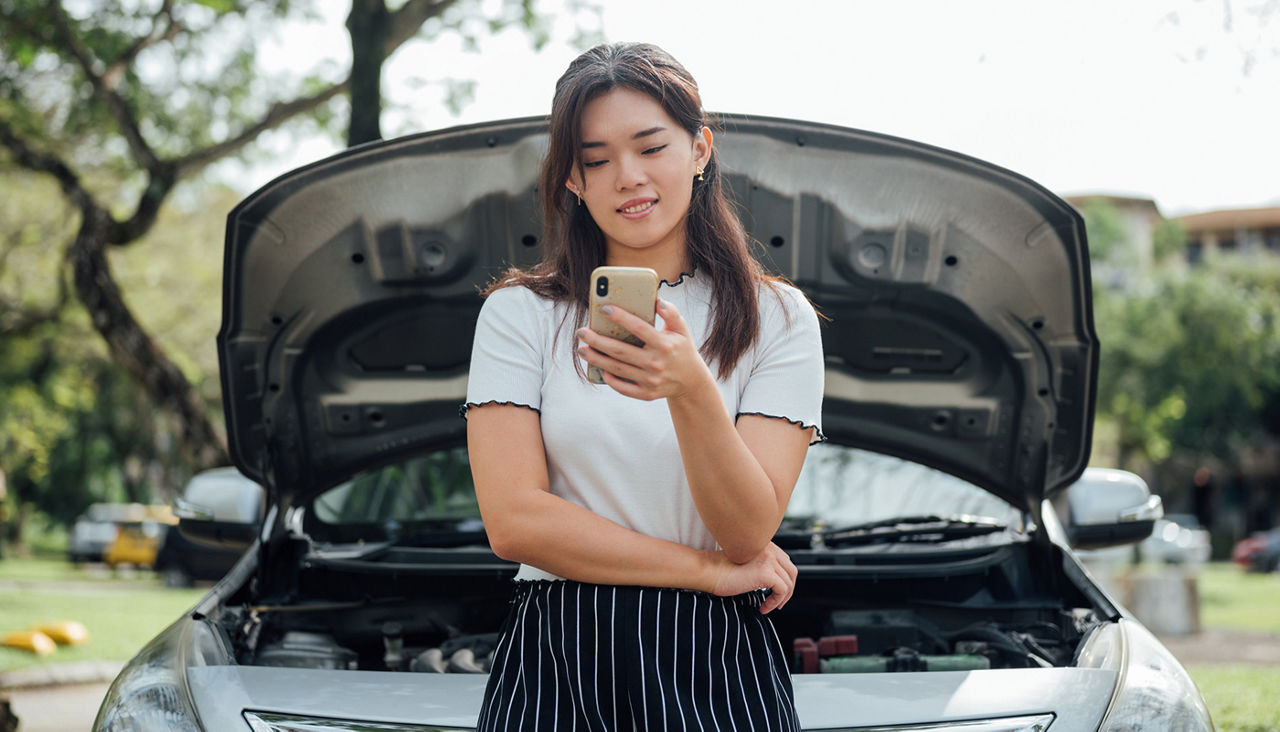 Smiling Young Asian woman waiting car assistant or someone for help with her broken carwhile standing near her car with open hood on the road side.