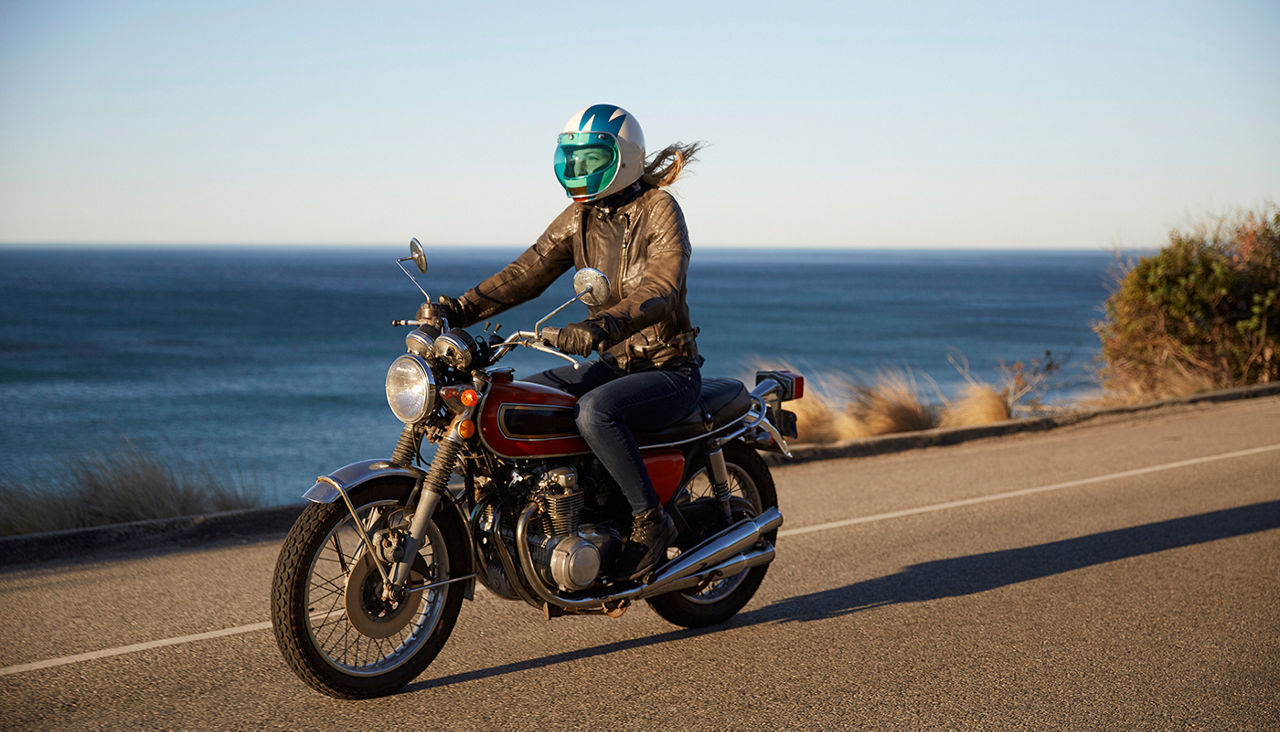 Female motorcyclist riding down a road along a coastline with the blue waters in the background.