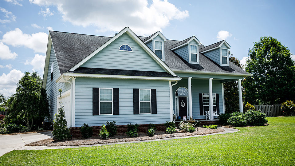 Pale blue house with siding on a large lot with traditional windows and shutters in a subdivision in the suburbs on a bright sunny blue sky day.