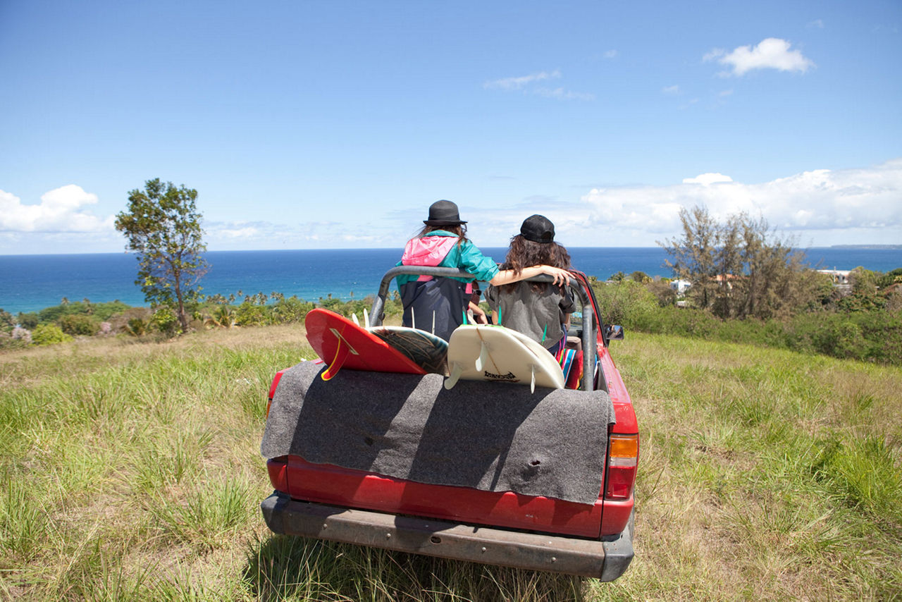 Two friends sit in their open truck looking out at the ocean why they get prepared to go surfing.