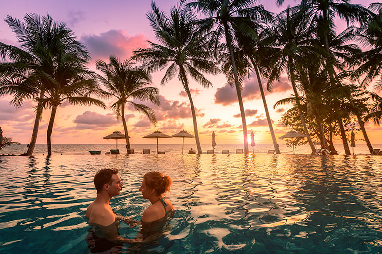 Couple at beach vacation holidays resort relaxing. in swimming pool with scenic tropical landscape at sunset, romantic summer honeymoon island destination, coconut palm tree near the sea