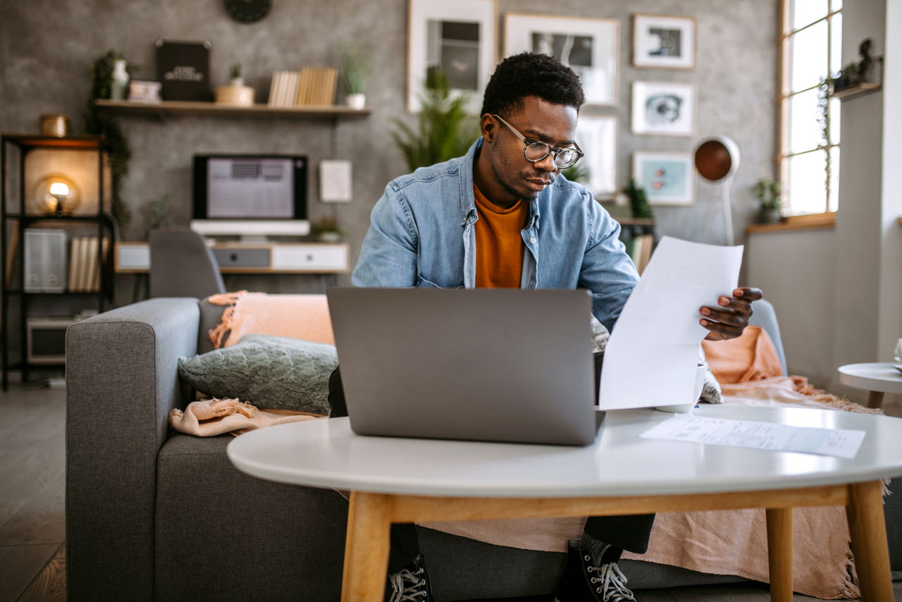 Young black man at home, sitting in sofa, checking financial documents, going through finances, paying bills online and planning investments.