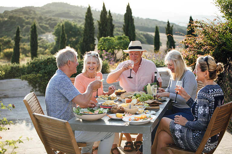 A group of mature friends are sitting around an outdoor dining table, eating and drinking. They are celebrating their holiday with a glass of champagne and enjoying each other's company. 