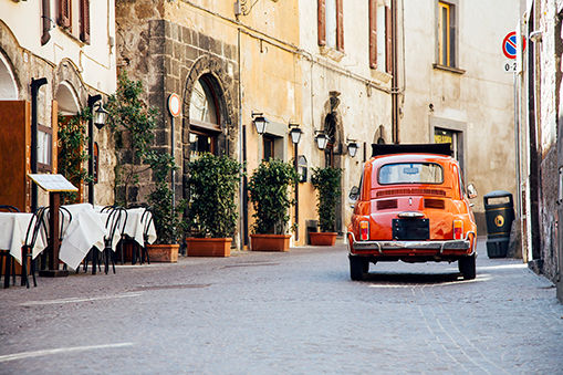 An orange buggie car is driving through the streets in a European town. 