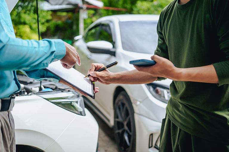 Clients engage with a car insurance agent beside a wrecked vehicle. They examine the policy details, assess the damage, and finalize paperwork related to the insurance claim and repairs.