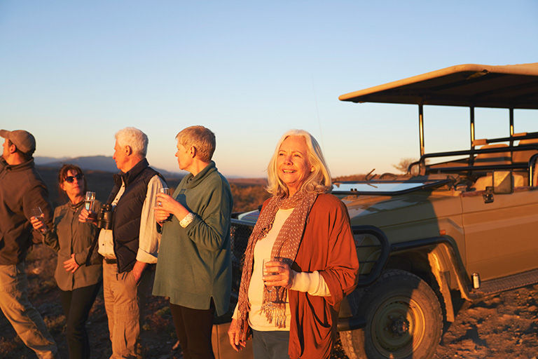 Carefree, active senior woman on safari drinking champagne outside off-road vehicle at sunset