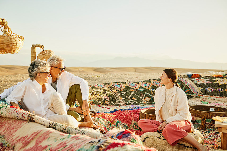 Wide shot of smiling senior parents relaxing with adult daughter while enjoying sunset at a luxury desert camp during family vacation exploring Morocco