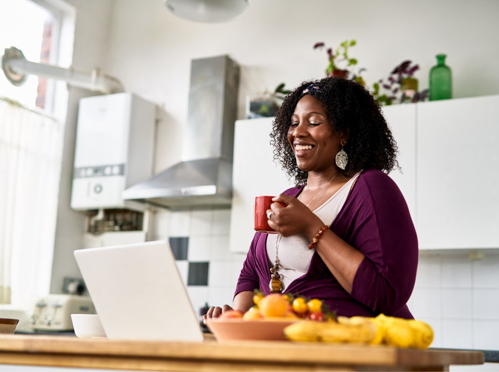 Woman in kitchen looking at her laptop and drinking a cup of coffee.