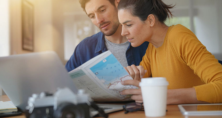 A couple reading travel articles on a laptop while looking at a map.