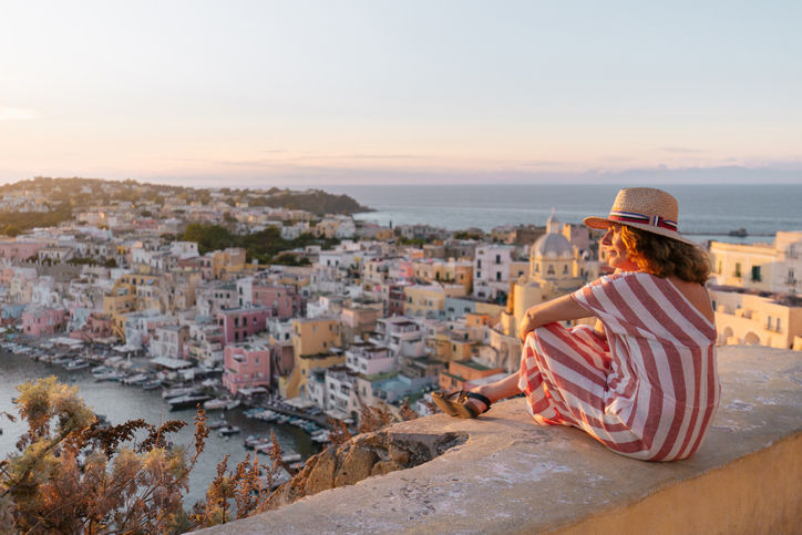 Side view of female traveler in striped dress and hat enjoying amazing seascape during sunset in Procida island.