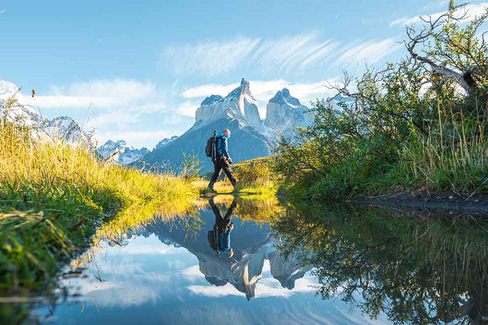 Man hiking across trail with mountains behind him, his reflection shines bright in the lake next to him.