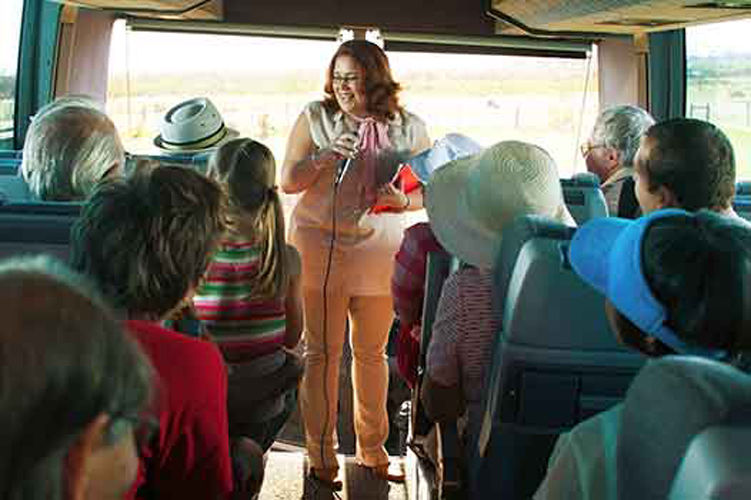 Women tour guide stands at the front of the bus speaking in the microphone.