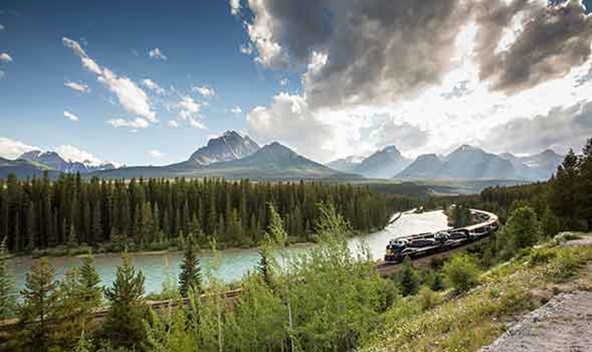 Train driving through the rocky mountains near a river and lush greenery.
