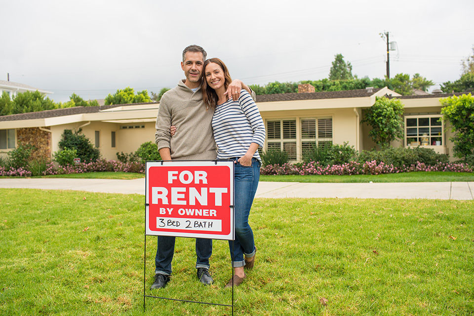 Couple behind for rent sign with house in background.