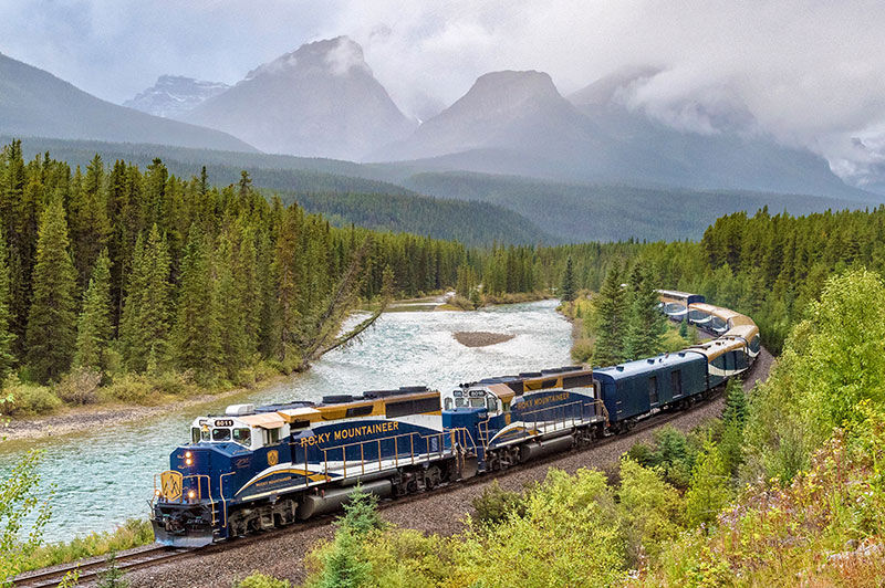 A train traveling along a river bed with red rocky mountains on the other bank.