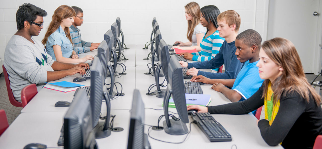 A multi-ethnic group of high school age students are sitting in a row at the computer lab and are doing research for an assignment online.