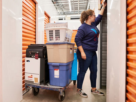 Woman opening the shutter of a self storage unit with a trolley full of goods and items in the aisle of self-storage building