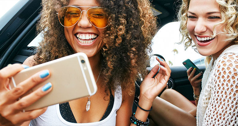 Two ladies look at a mobile phone from the edge of their car.