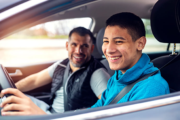 A teenage boy sitting behind the steering wheel of a car and listening to his fathers instructions as he drives.