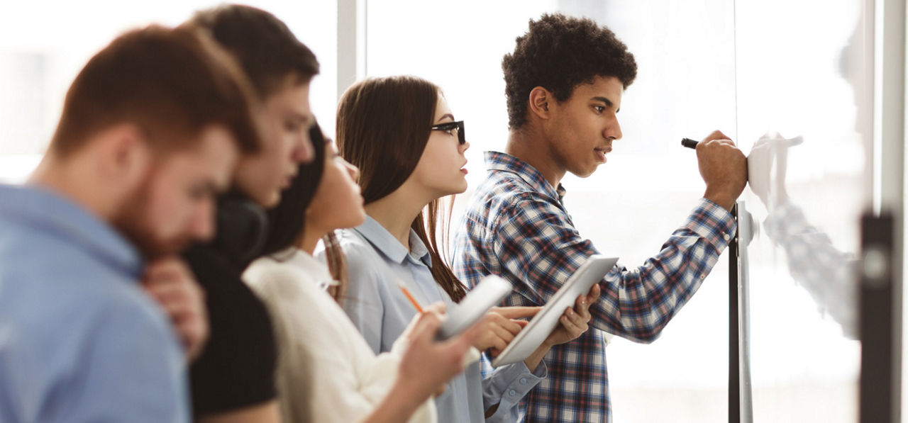 Male high school pupil writing on whiteboard in class, classmates checking task answers