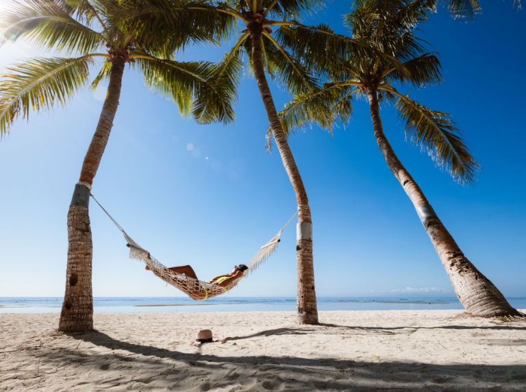 Person laying on a beach front hammock surrounded by palm trees.