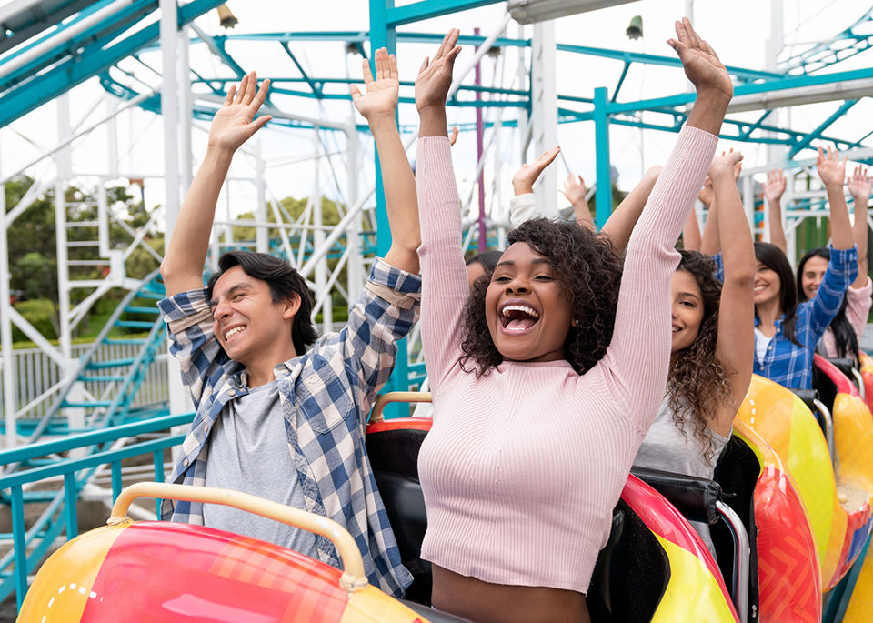 Happy group of friends having fun in an amusement park riding on a rollercoaster with arms up and screaming.