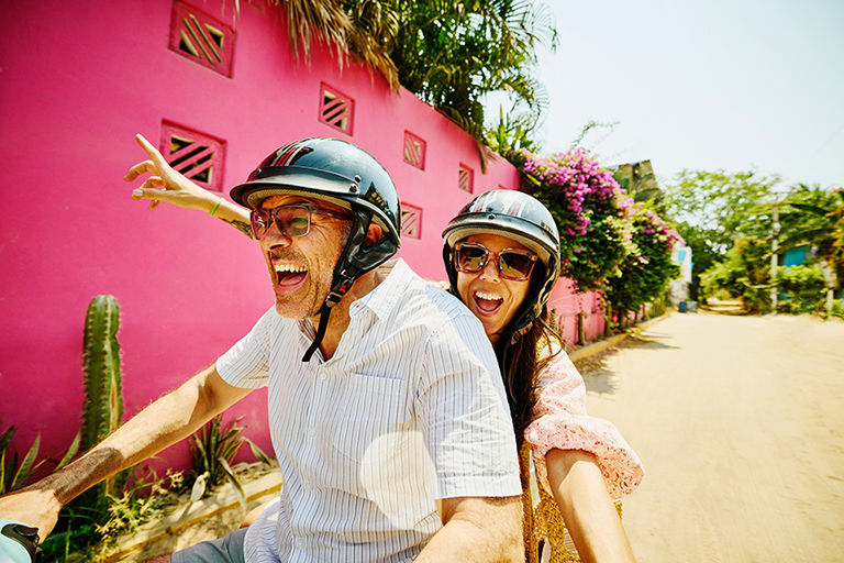 Mature couple with helmets on laughing while riding on a moped on vacation.