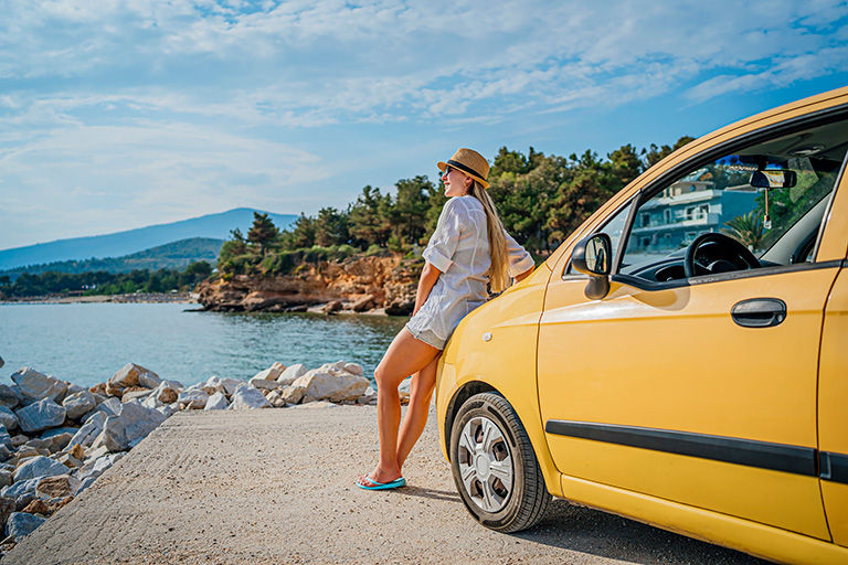 Woman on road trip leaning on the front of her rental car looking at the water.