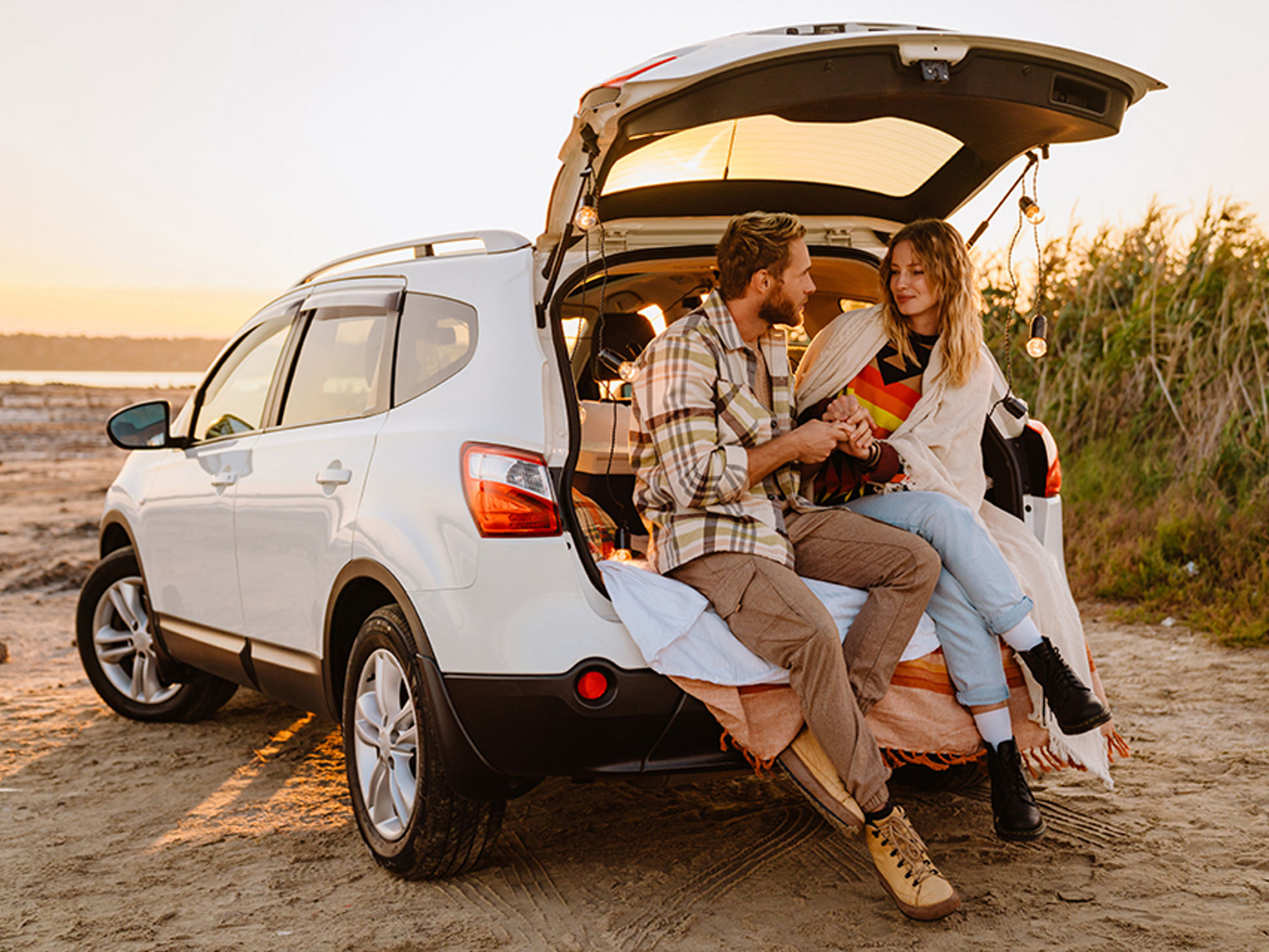 Happy young couple smiling and sitting in by car trunk near beach.