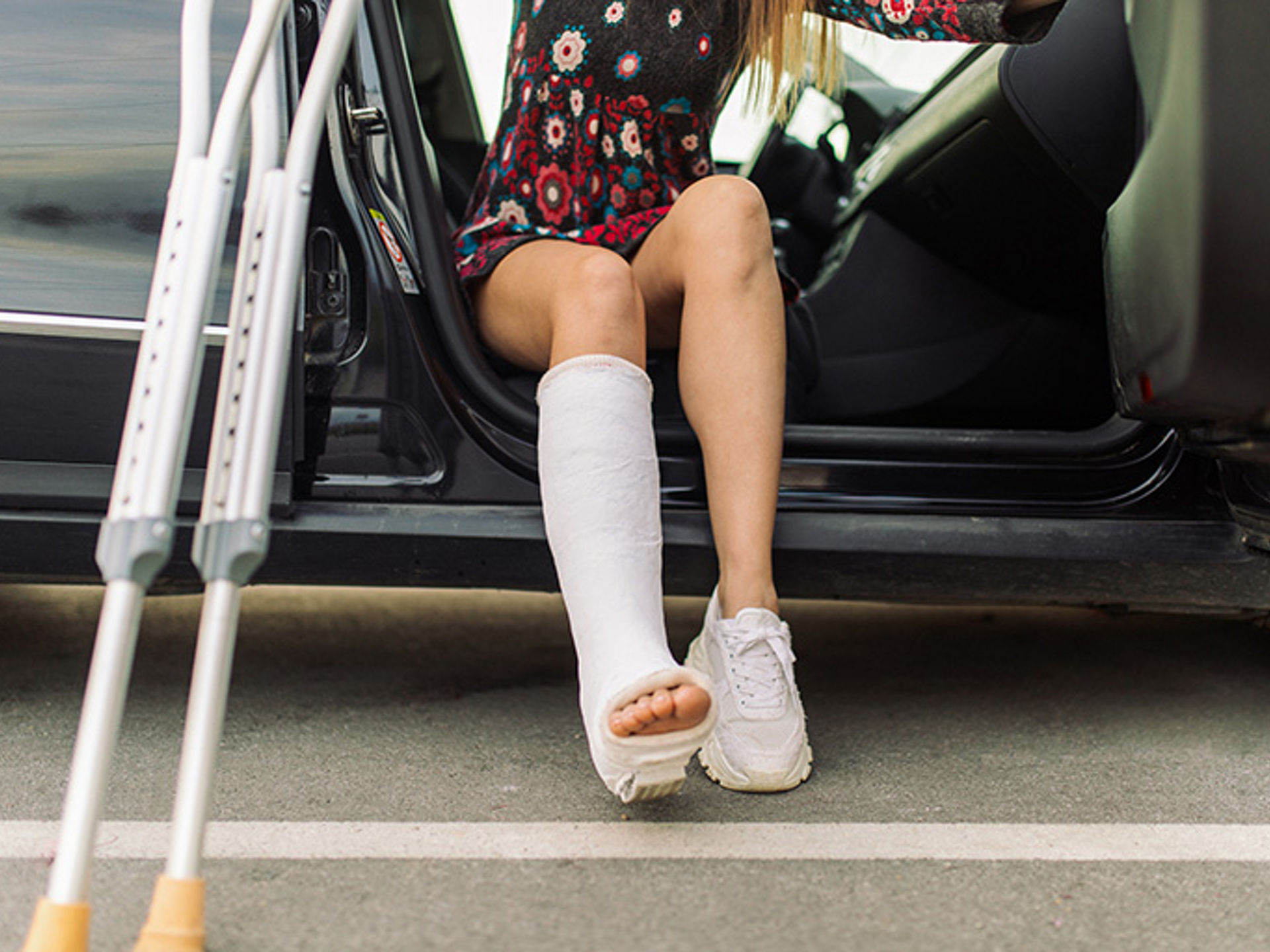 A mid adult woman with a cast on her leg in a beautiful floral long sleeved dress, getting out of car at parking lot downtown, holding a crutch